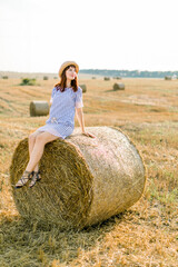 Naklejka na ściany i meble Countryside landscape of farm wheat field at summer sunset. Pretty young happy Caucasian woman in straw hat and blue striped dress sitting on hay bale and enjoying warm evening and vacations