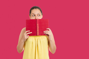 Back to school. Portrait of a student who covers her face with a book and looks to the right. Copy space. Isolated on a red background. Concept of education and reading