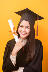 Portrait of happy Beautiful woman in graduation gown is holding education certificate on yellow...