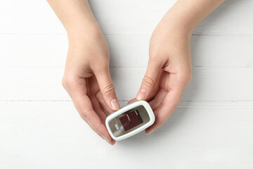 Woman holding fingertip pulse oximeter on white wooden background, top view