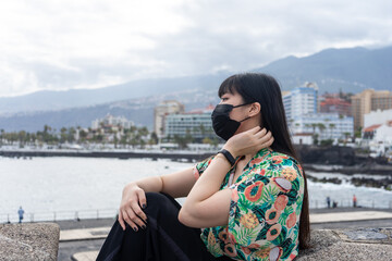 Fototapeta na wymiar Young Asian tourist sitting watching the sea. She is touching her hair. Wear a floral shirt and mask. Sightseeing in Puerto De la Cruz, Tenerife.