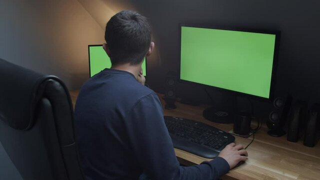 Young man sitting at his desk with two green screen monitors over shoulder shot