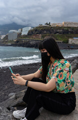 Fototapeta na wymiar Young Asian tourist chatting with her smartphone. It is located on a black sand beach, in Puerto De La Cruz in Tenerife. She wears a floral shirt and mask.