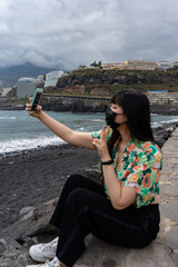 Fototapeta na wymiar Young Asian tourist taking a selfie with her smartphone. It is located on a black sand beach, in Puerto De La Cruz in Tenerife. She wears a floral shirt and mask.