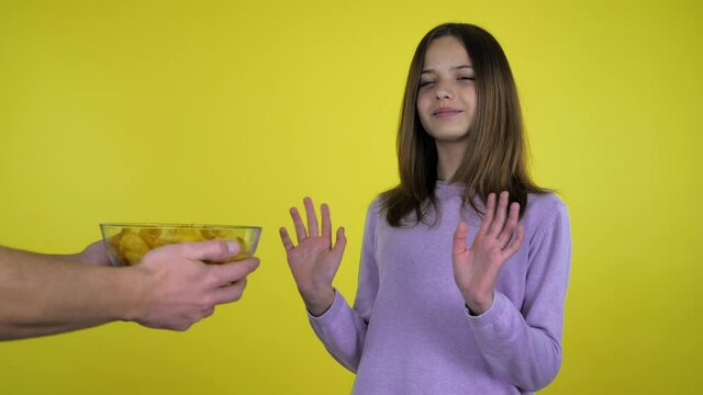 Teen girl in a pink sweater refuses potato chips in a glass bowl shaking hands and head on a yellow background with copy space. Place for text or product. 4K slow motion footage