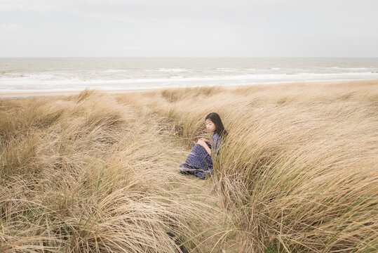 Girl in classic blue dress sitting and contemplating in grass dunes near ocean