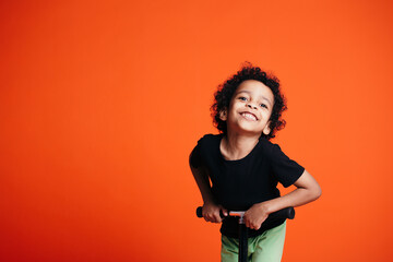 A curly-haired boy in a black T-shirt rides a scooter and playfully leans forward towards the...