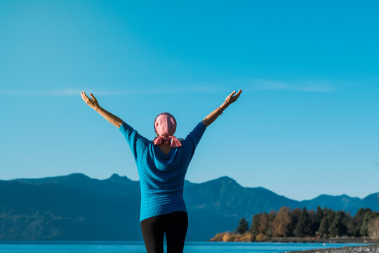 Woman with cancer wear a pink headscarf with her arms open looking the landscape