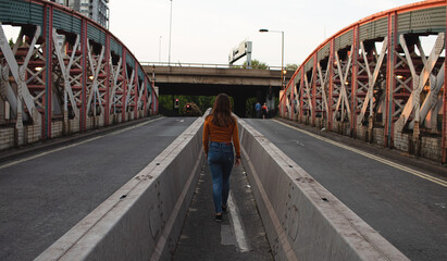 Attractive young woman walking on the middle of a road and crossing a bridge while wearing a face...