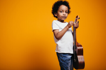 A little black boy holds a ukulele guitar in his hands and pulls up the strings so that they sound...