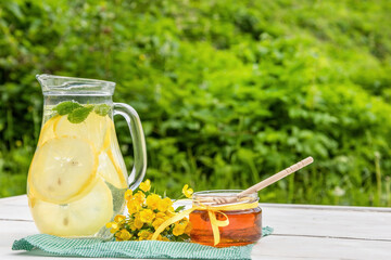 A jug of mint lemonade and a jar of honey on the table in the summer green garden.