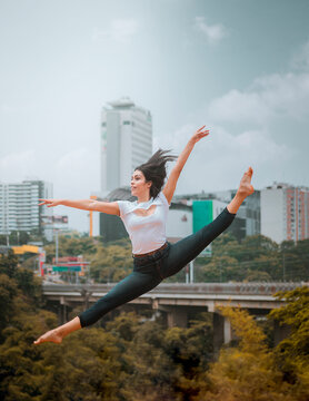 Full body energetic barefoot woman in casual clothes jumping and doing splits while dancing against contemporary city and cloudy sky
