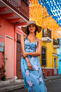 Calm young female in stylish blue summer dress and hat looking at camera while standing near aged building with colorful walls on city street