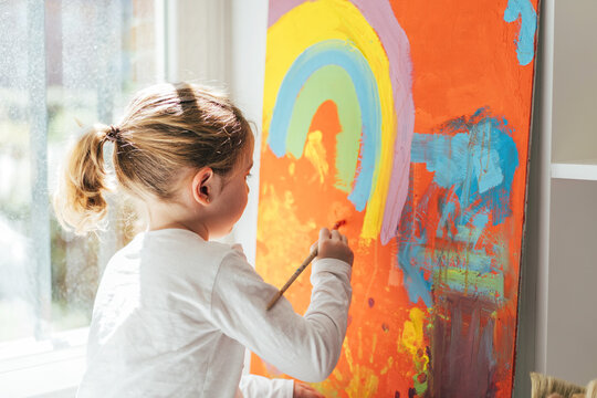 Creative blond girl in casual clothes sitting on window sill against window and painting with paintbrush large multi colored rainbow on orange canvas