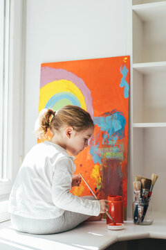 Creative blond girl in casual clothes sitting on window sill against window and painting with paintbrush large multi colored rainbow on orange canvas
