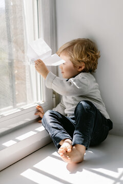 Side view of blonde little child in casual clothes playing with paper airplane while sitting barefoot on window sill on sunny day