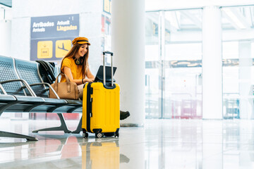 Side view of serious female traveler in stylish clothes sitting on bench with legs crossed with luggage and spending free time using laptop while waiting for boarding in airport