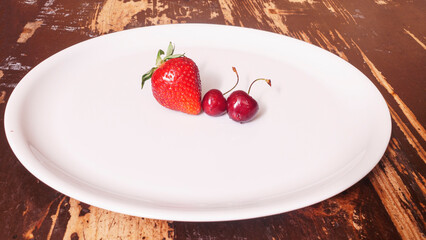Top view of red strawberry and two sweet red cherries in the white plate on a wooden background. Space for text. Design concept.