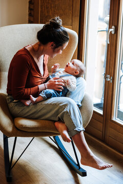 Full body barefoot woman in casual clothes smiling and using baby talk with adorable infant while sitting on comfortable chair near window at home