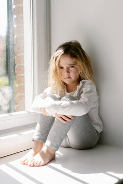 Annoyed little girl with curly hairs wearing gray cozy pajamas looking at the camera with dissatisfaction while sitting on sill with crossed arms