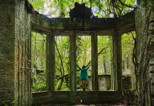 Back view of anonymous blond female with long hair standing with arms raised while touching ancient columns in quiet abandoned place with trees in sunlight