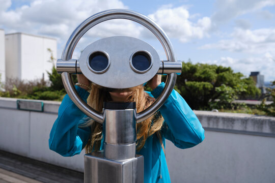 Anonymous blond female in blue coat looking through street coin operated binocular while standing on wooden pavement near concrete fence on sunny day