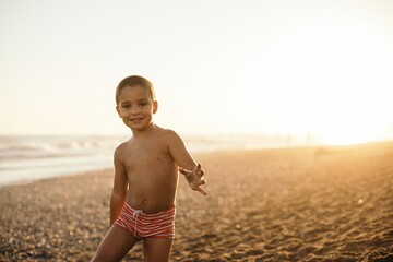 Happy shirtless boy smiling and looking at camera while standing on sandy beach during sunset