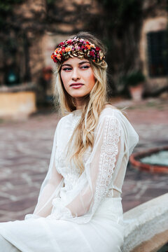 Pretty young woman in translucent white dress and floral wreath looking away while sitting on blurred background of yard on wedding day