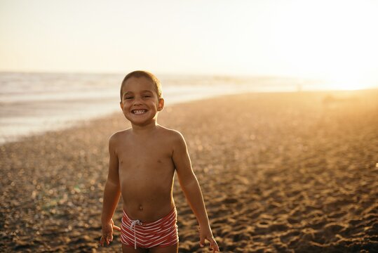 Happy shirtless boy smiling and looking at camera while standing on sandy beach during sunset