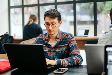 Young concentrated male in casual outfit and eyeglasses sitting at desk and typing on laptop while working on project in creative workspace