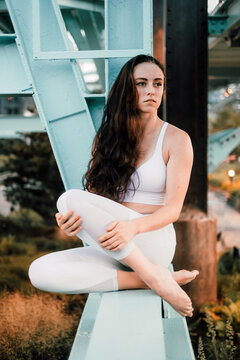 Dreamy female in sportswear sitting on metal balk of urban bridge while enjoying cityscape and looking away