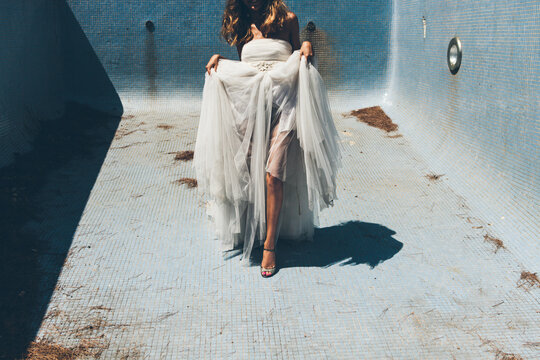 Happy young anonymous bride  in elegant white wedding dress dancing sensually in empty dirty pool on sunny day during romantic party