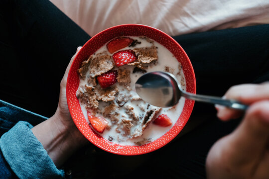 From Above Crop Female In Casual Clothes Holding Metal Spoon And Red Bowl With Pieces Of Strawberry And Oatmeal Poured With Milk While Sitting On Bed In Light Bedroom