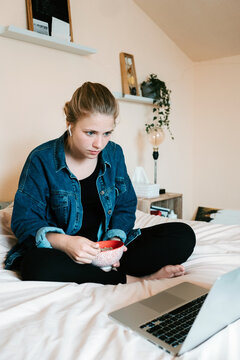 Stunned pensive barefoot female in true wireless earphones and casual clothes with bowl of tasty food focusing on screen with interest while sitting with legs crossed on soft bed and using netbook in cozy apartment
