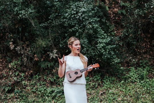 Cheerful bride playing ukulele near bushes
