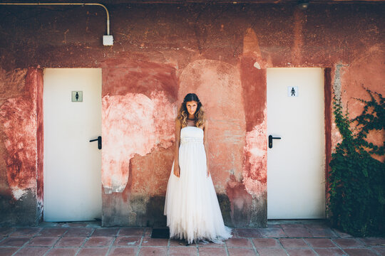 Charming young bride in elegant white wedding dress standing near red shabby wall near toilet doors and looking at camera