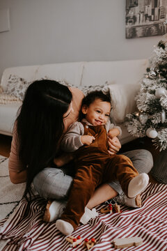 From above view of cheerful young mother in casual clothes hugging cute smiling little son while sitting with crossed legs on carpet on floor near decorated Christmas tree