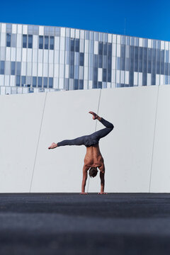 Back view of unrecognizable shirtless sportsman performing handstand with one hand while doing exercises against wall and blue sky on rooftop of modern building