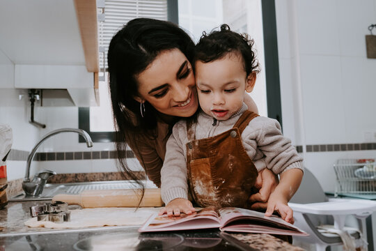 Cheerful mother smiling and hugging son while reading recipe book on table during cooking delicious cookies in light kitchen at home