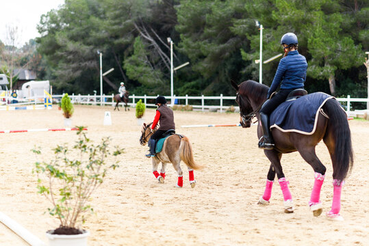 Back view of boy on roan pony and teen girl on brown horse riding horses on dressage arena during lesson in equestrian school
