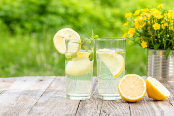 Two glasses of lemonade with lemon and mint for two on a background of greenery and a bouquet of field yellow flowers.