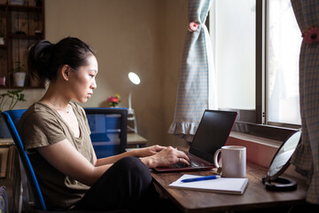 Side view of Asian female freelancer in casual t shirt and eyeglasses sitting at table and browsing computer while working on project online at home