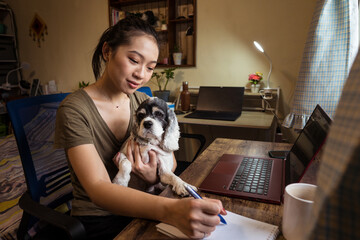 Side view of focused female freelancer sitting on chair taking notes on notepad while holding a dog working remotely from home