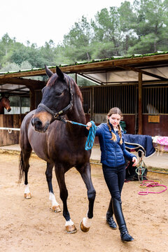 Full body teen jockey pulling reins of brown horse while walking on sandy ground of dressage arena during lesson in equestrian school