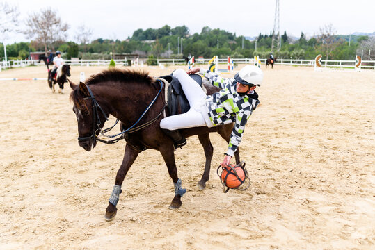 Teen jockey playing with ball on sandy ground from back of brown horse during lesson in equestrian school