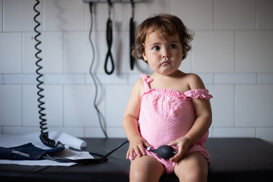 Curious adorable little girl in casual pink dress sitting on bench in medical room and playing with tonometer while visiting doctor