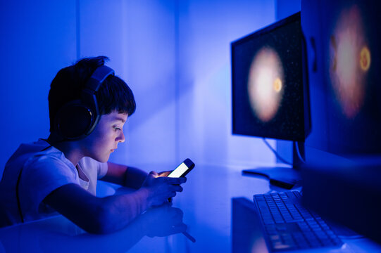 Side view of serious boy in headphones surfing internet on cellphone while sitting in front of desktop computer in room with blue light