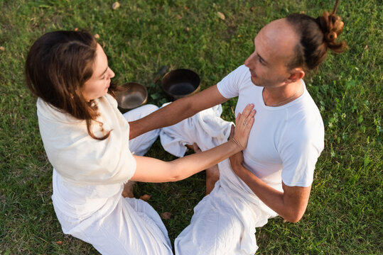 From above of calm couple in white clothes sitting opposite each other in lotus position on grass and keeping hands on stomachs and on each others hearts while meditating outdoors with singing bowls in park