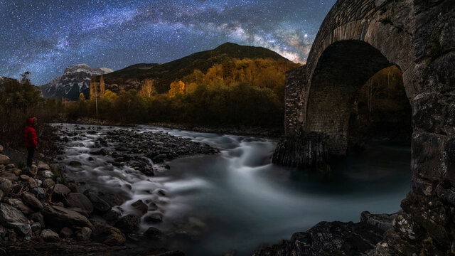 Side view of explorer standing on stones in dark rocky terrain with green trees and looking at mountain stream flowing through rocky cliff and stone old bridge on background of mountains in starry night