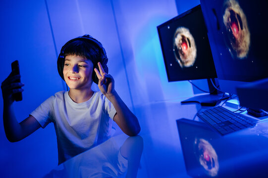 Content male child in headphones taking selfie on cellphone while sitting with thumb up at table with desktop computer in room